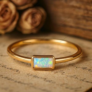 Könnte beinhalten: Ein goldener Ring mit einem rechteckigen Opalstein. Der Opal zeigt eine Mischung aus Farben, darunter Blau, Grün und Weiß. Der Ring liegt auf einer hellbraunen Oberfläche mit Schrift.