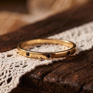 May include: A gold band ring with small black gemstones set into the band. The ring is displayed on a piece of white lace over a dark wooden surface. The ring is a simple, elegant design.