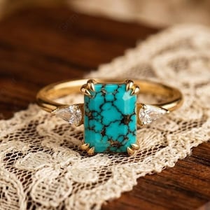 May include: A gold ring featuring a rectangular turquoise stone with black veining. The ring has two small, clear, triangular accent stones on either side of the turquoise center stone. The ring is set on a wooden surface with a lace trim.