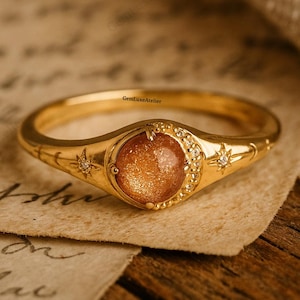 Könnte beinhalten: Goldener Ring mit einem runden, orange-braunen Edelstein in der Mitte. Der Ring hat Stern- und Halbmond-Designs mit kleinen, klaren Akzentsteinen. Der Ring liegt auf einem Stück gealtertem Papier.