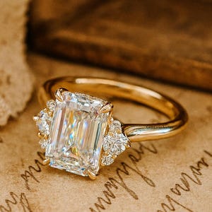 May include: A gold ring featuring a large, rectangular-cut diamond as the centerpiece, flanked by smaller accent stones. The ring's band is a polished gold, and the setting showcases the brilliance of the diamonds. The ring is set on a textured, tan surface.