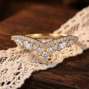 Pode incluir: Um anel dourado com um design em forma de V, cravejado com pedras preciosas claras e brilhantes. O anel é adornado com pedras menores na parte superior e pedras maiores com corte marquesa no centro. O anel repousa sobre uma fita de renda creme.