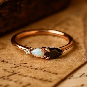 Può includere: Un anello in oro rosa con un'opale bianca, un onice nero e un piccolo diamante. Le pietre preziose sono incastonate in un design delicato e minimalista. L'anello è esposto su uno sfondo di carta vintage, evidenziando il suo fascino elegante e senza tempo.