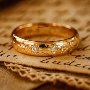 Puede incluir: Un anillo de oro con detalles de estrellas y diamantes. El anillo presenta diseños en forma de estrella, cada uno con un diamante central. El anillo está sobre un fondo de papel envejecido con escritura manuscrita.