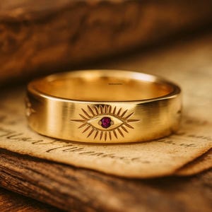May include: A gold ring with an engraved eye design and a small, round, red gemstone in the center. The ring is set on a piece of aged paper, creating a vintage aesthetic. The ring's band is wide and has a brushed finish.