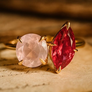 May include: A gold ring featuring two gemstones: a round, light pink rose quartz and a pear-shaped, deep red ruby. The gemstones are held in place by gold prongs.