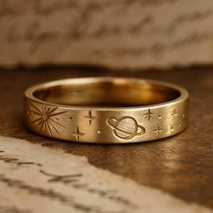 Puede incluir: Un anillo dorado con un diseño celestial grabado. El anillo presenta un sol, estrellas y un planeta con anillos. El anillo es de estilo banda y tiene un acabado cepillado.