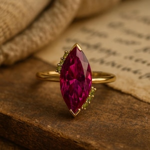 May include: A gold ring with a large, marquise-cut, deep pink gemstone. The ring is adorned with small, green gemstones on either side of the main stone. The ring is set on a wooden surface.