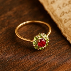 May include: A gold ring with a central red gemstone, surrounded by green gemstones in a floral design. The ring is set on a wooden surface, with a piece of parchment in the background. The ring is from GemLuxeAtelier.