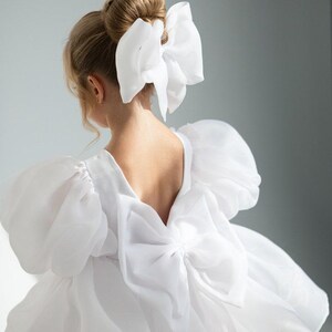 May include: A white dress with a large bow on the back and a matching bow in the hair. The dress has puffy sleeves and a V-neck back. The image is a close-up of the back of the dress and the person's hair.