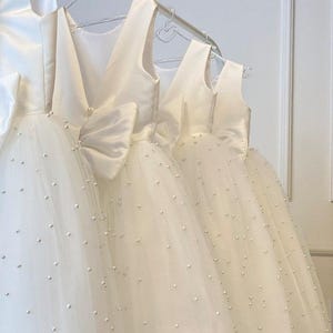 May include: Several ivory-colored dresses hanging on a rack. The dresses have a V-neck design and a large bow at the back. The skirts are made of tulle and are adorned with small, scattered pearls. The dresses are likely for a special occasion.