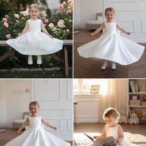 May include: A little girl in a white satin dress with a full skirt. The sleeveless dress has a simple, elegant design. The girl is smiling and twirling in the dress, showcasing its flowing movement. The dress is suitable for special occasions.