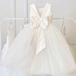 May include: A white sleeveless dress with a large bow at the waist. The dress has a V-neck and a full, layered skirt adorned with small, scattered pearls. The dress is hanging on a white rack with other dresses.