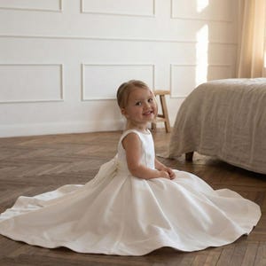 May include: A young child wearing a white satin dress with a full skirt and a bow in the back. The dress has a sleeveless design and a round neckline. The child is sitting on a wooden floor in a room with white walls.