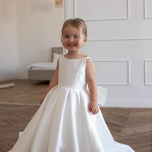 May include: A young child wearing a white satin dress with a full skirt and a bow in the back. The dress has a simple, elegant design. The child is smiling and standing in a room with light-colored walls and a wooden floor.