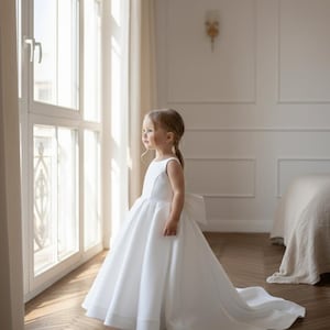 May include: A young child wearing a white sleeveless dress with a full skirt and a long train. The dress features a large bow on the back. The child is standing near a window, looking out. The room has light-colored walls and a wooden floor.