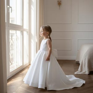 May include: A young girl in a white sleeveless dress with a full skirt and a long train. The dress features a large bow on the back. The girl is standing near a window, looking out. The dress is likely for a special occasion.