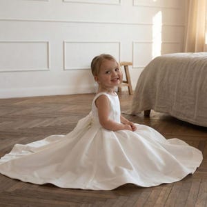 May include: A young child wearing a white sleeveless dress with a full skirt and a bow in the back. The dress is floor-length and the child is sitting on a wooden floor. The background includes a white wall and a bed.