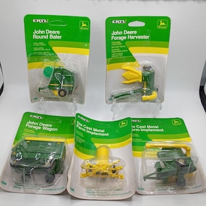 May include: Five John Deere die-cast metal farm implement toys in their original packaging. The toys include a round baler, forage harvester, forage wagon, and other farm implements. The packaging is green and yellow.