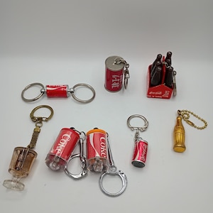 May include: A collection of Coca-Cola themed keychains. The keychains include miniature Coke cans, bottles, and a six-pack of bottles. The keychains are red, silver, and gold in color. Some keychains have the word "Coke" printed on them.