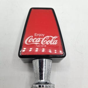 May include: A vintage Coca-Cola tap handle. The handle has a red top with the Coca-Cola logo and the words "Enjoy Coca-Cola". The handle is black and has a silver-colored base.