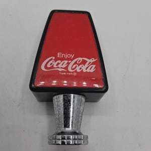 May include: A vintage Coca-Cola tap handle with a red and black design. The handle features the Coca-Cola logo in white, with the words "Enjoy" above and "Trade mark" below.