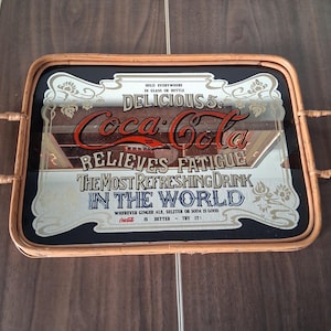 May include: Vintage Coca-Cola serving tray with a mirrored surface and a bamboo frame. The tray features the Coca-Cola logo in red and white, along with the text "Delicious 5¢" and "Relieves Fatigue". The tray has handles on each side.