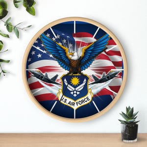 May include: A round wall clock with a wooden frame. The clock face features a patriotic design with an American flag background, an eagle with outstretched wings, and two fighter jets. The words "U.S. Air Force" are printed on the clock face.