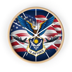 Op de afbeelding: Een ronde wandklok met een houten frame. De wijzerplaat heeft een patriottisch ontwerp met een Amerikaanse vlag, een adelaar en twee straaljagers. De woorden "U.S. AIR FORCE" staan op de wijzerplaat.