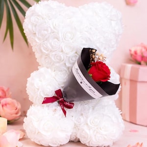 以下が含まれることがあります： 白いバラのクマが赤いバラの花束を持っています。クマは小さな白いバラでできています。花束は黒い紙で包まれ、赤いリボンで結ばれています。背景はソフトピンクです。
