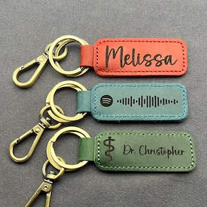 Puede incluir: Tres llaveros con herrajes dorados y etiquetas de cuero. Las etiquetas son rojas, verde azulado y verdes. La etiqueta roja dice "Melissa", la etiqueta verde azulado tiene un código de Spotify y la etiqueta verde dice "Dr. Christopher".