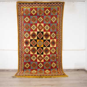 Può includere: Un tappeto rettangolare con un motivo geometrico in rosso, giallo e blu. Il design centrale presenta quadrati neri e gialli. Il tappeto ha un bordo rosso e una frangia gialla. È esposto contro un muro bianco, mostrando il suo design intricato.