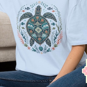 May include: A light blue t-shirt featuring a circular graphic of a sea turtle surrounded by coral, small fish, and seaweed. The turtle shell has intricate patterns in shades of teal, orange, and white. The text Light Blue is visible.