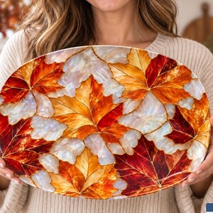 Puede incluir: Una bandeja de servicio ovalada con un vibrante patrón de mosaico de hojas de otoño en tonos rojo intenso, naranja quemado y amarillo dorado, intercalado con segmentos blancos iridiscentes y nacarados que crean un efecto brillante.