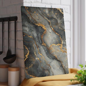 May include: A rectangular glass cutting board featuring a marbled pattern in shades of charcoal gray, slate, and white, accented with metallic gold veins and scattered gold flecks across the surface, creating a fluid and layered visual effect.