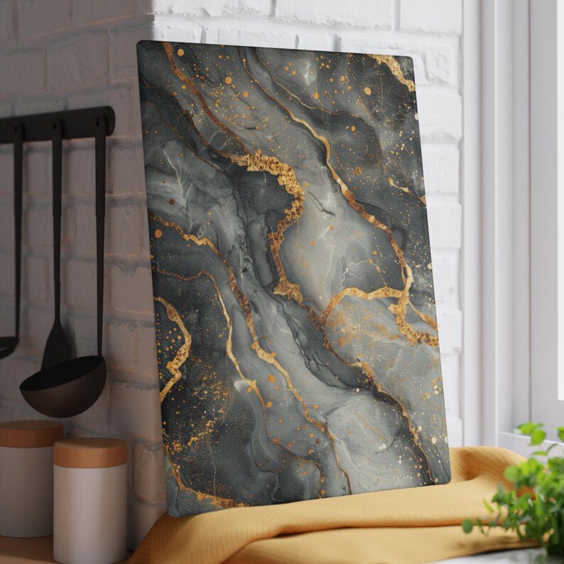 May include: A rectangular glass cutting board featuring a marbled pattern in shades of charcoal gray, slate, and white, accented with metallic gold veins and scattered gold flecks across the surface, creating a fluid and layered visual effect.