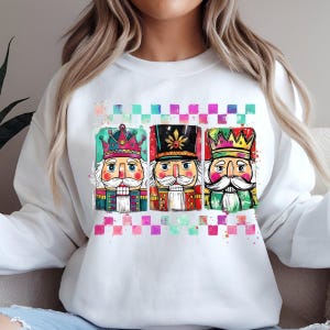 Może przedstawiać: Biała bluza z kolorową grafiką przedstawiającą trzy postacie dziadka do orzechów. Każdy dziadek do orzechów ma unikalny design, w tym korony i szczegółowe mundury. Projekt umieszczono na kratkowanym tle z akcentami akwarelowymi.