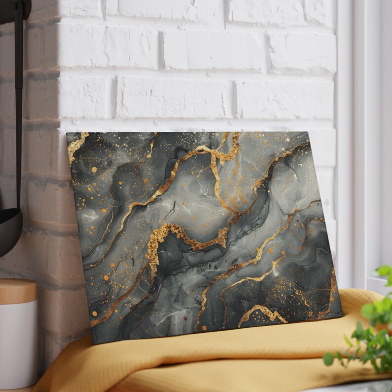 May include: A rectangular glass cutting board featuring a marbled design with swirling shades of charcoal gray, black, and white, accented by prominent veins of metallic gold and scattered gold flecks across the entire surface.