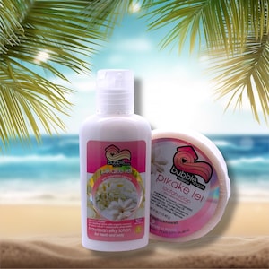 Peut inclure: Une bouteille blanche de lotion soyeuse hawaïenne et un savon loofah rond avec le texte "pikake lei" sur une étiquette rose. La lotion est pour les mains et le corps. Le fond montre une scène de plage avec des feuilles de palmier.