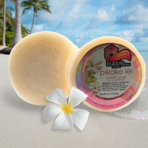 Peut inclure: Deux savons loofah ronds et jaune clair "Pikake Lei" avec une fleur blanche sur une plage de sable. Un savon a une étiquette avec du texte et un logo. L'étiquette indique "Bubble Shack Hawaii" et répertorie les ingrédients. Chaque savon pèse 113,40 grammes.