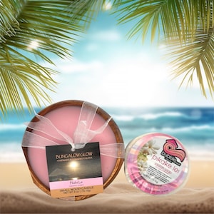 Peut inclure: Une bougie rose dans un bol en bois avec un ruban blanc et une étiquette "BUNGALOW GLOW". À côté, un savon rond "pikake lei". La bougie pèse 170.10g. En arrière-plan, une scène de plage.