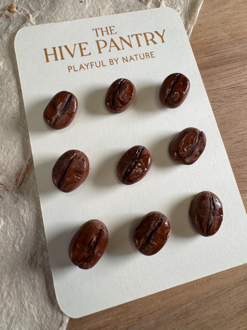 May include: A card with the text "THE HIVE PANTRY PLAYFUL BY NATURE" displays nine glossy, brown coffee bean-shaped objects. The beans are arranged on a white card, suggesting a decorative or craft item related to coffee or kitchen decor.
