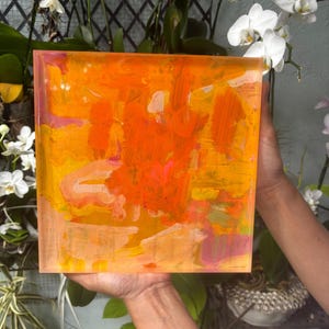 May include: Square abstract art piece with vibrant orange, yellow, and pink brushstrokes. The artwork has a translucent appearance, with visible layers of paint. The piece is held up in front of a background of white orchids and greenery.