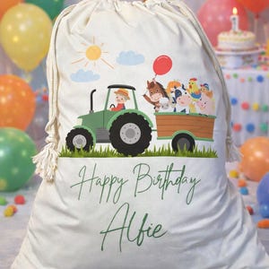 May include: A cream-coloured drawstring bag featuring a green tractor pulling a wooden trailer filled with cartoon farm animals, including a horse, cow, chicken, and pig. The text Happy Birthday Alfie is printed in green script below the tractor.