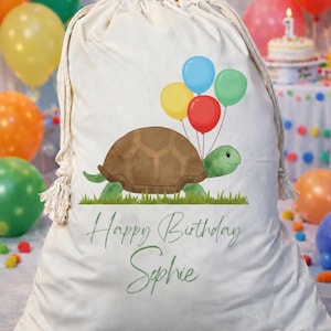 May include: A cream-coloured drawstring bag featuring a watercolour illustration of a green turtle holding four colourful balloons in red, yellow, blue, and green. Below the turtle, the text reads Happy Birthday Sophie in a green cursive font.