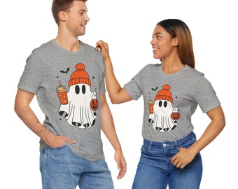 May include: Two gray t-shirts with a cartoon ghost design. The ghost wears an orange beanie and holds a pumpkin and a drink. A black bat graphic is above the ghost. The shirts are paired with blue jeans.