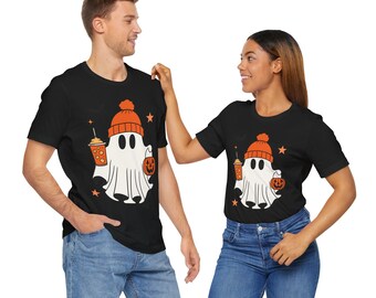 May include: Black t-shirts featuring a cartoon ghost wearing an orange beanie and holding a pumpkin and a coffee cup. The ghost has a white body and black eyes. The shirts are worn by two people.