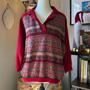 May include: A red long-sleeved sweater with a collared neckline and a button closure. The sweater features a patterned design in shades of red, brown, and blue. The pattern includes geometric shapes and horizontal stripes. The sweater is displayed on a mannequin.