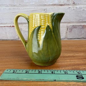May include: A ceramic jug with a corn-on-the-cob design. The jug features a yellow corn cob body with green husk accents and a curved handle. The jug is approximately 15 cm tall.