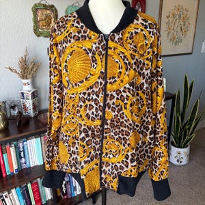 May include: A zip-up bomber jacket with a striking leopard print design, featuring gold baroque accents and a black collar and cuffs. The jacket is displayed on a mannequin, showcasing its full length and design.